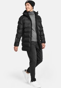 Chaqueta de plumas negra con capucha, con un diseño acolchado y cierre de cremallera. Lleva puesta una camiseta de cuello alto gris y la ha combinado con unos jeans oscuros y zapatillas blancas.