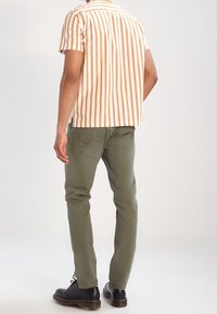 Košile s krátkým rukávem s vertikálními oranžovými a bílými pruhy, v kombinaci s olivově zelenými kalhotami a černými botami, viděná ze zadu.