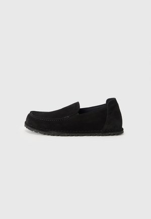 Mocassin noir en daim à enfiler avec des détails cousus et une semelle en caoutchouc flexible, présenté de profil sur un fond clair uni.