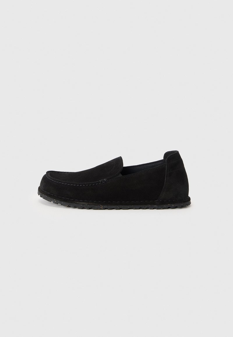 Mocassin noir en daim à enfiler avec des détails cousus et une semelle en caoutchouc flexible, présenté de profil sur un fond clair uni.
