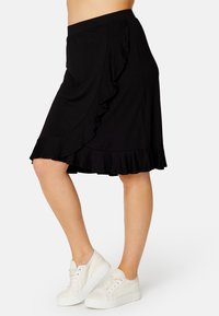 Gonna nera lunga fino al ginocchio con orlo arricciato e drappeggio laterale. Realizzata in tessuto morbido, abbinata a sneakers bianche.