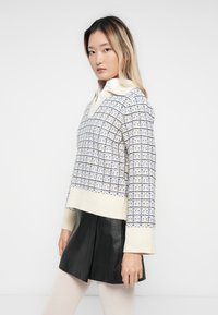Giovane donna con capelli lunghi che indossa un maglione lavorato a maglia con trama sopra una camicia bianca con colletto, una gonna di pelle nera e collant bianchi, su uno sfondo neutro.