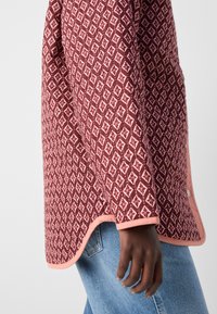 Långärmad jacka i vinröd med diamantmönster, med rosa detaljer och slät textur. Bärs över ljusblå jeans och visar en sidovy.