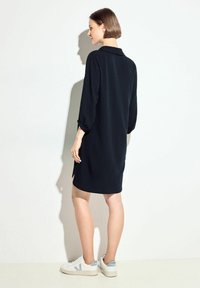Schwarzes knielanges Kleid mit einem Kragen und umgeschlagenen Ärmeln. Getragen mit weißen Sneakers mit hellblauen Akzenten. Texturiertes Material.