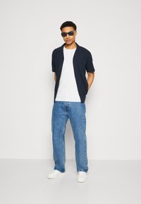 Camisa de botón navy de manga corta, camiseta blanca debajo, jeans azules y zapatillas blancas. El conjunto se completa con gafas de sol negras.