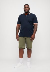 Navy polo shirt with red and white striped collar, olive green shorts, and white sneakers. Smooth fabric with gradual hem on shorts.