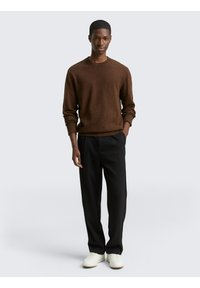 Brauner Strickpullover mit Rundhalsausschnitt und gerippten Bündchen, kombiniert mit schwarzen, plissierten Hosen und weißen Sneakers vor einem neutralen Hintergrund.