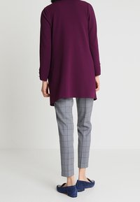 Kobieta stojąca tyłem, ubrana w długi fioletowy żakiet, szare spodnie w kratę oraz niebieskie zamszowe mokasyny na białym tle.