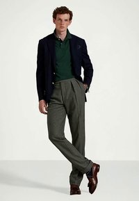 Άνδρας που φοράει σκούρο πράσινο πόλο μπλουζάκι, ναυτικό σακάκι, γκρι πλισέ παντελόνι, καφέ δερμάτινα παπούτσια, στέκεται με το ένα πόδι σταυρωμένο, με το χέρι στην τσέπη.