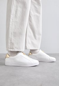 Baskets en cuir blanc avec accents dorés et fermeture éclair. Tige texturée, semelle plate et design minimaliste. Le modèle porte un pantalon clair.