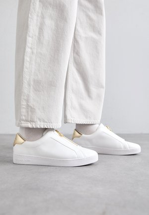 Hvite lær sneakers med gullakenter og glidelås. Teksturert overdel, flat såle og minimalistisk design. Modellen har på seg lyse bukser.