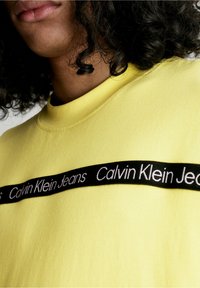 Žlutá bavlněná mikina se černým pruhem s nápisem "Calvin Klein Jeans" bílým písmem přes hrudník; kulatý výstřih a volný střih.