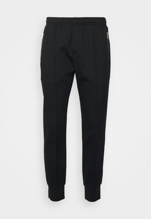 Black jogger pants with an elastic waistband, tapered fit, and zippered side pockets. Made of smooth fabric with ribbed cuffs.