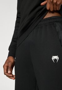 Μαύρο αθλητικό παντελόνι με ελαστική μέση, που διαθέτει λευκό λογότυπο φιδιού στον μηρό και υφασμάτινες διακοσμήσεις. Λείο, ελαστικό ύφασμα.