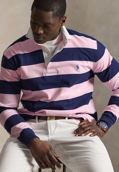 Mann trägt navy- und pink gestreiftes Rugbyshirt, weiße Hose, braunen Gürtel und Armbanduhr, sitzt und schaut nach unten.