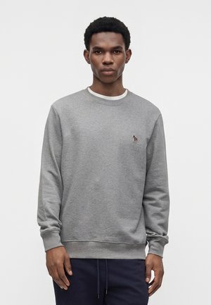 Jeune homme portant un sweat-shirt gris uni avec un petit logo zèbre brodé sur la poitrine gauche, associé à un pantalon foncé.