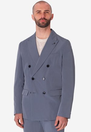 Uomo con barba corta che indossa una giacca doppiopetto azzurro chiaro e camicia bianca su uno sfondo uniforme.
