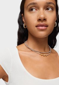 Collana multistrato argentata con dettagli in oro e legno, posizionata attorno al collo, indossata con un top bianco testurizzato e orecchini a cerchio dorati.