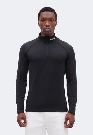 Άνδρας στέκεται, φοράει μαύρο μακρυμάνικο μπλουζάκι με φερμουάρ και το κείμενο "BORG" στον γιακά, και άσπρο παντελόνι μπροστά σε απλό φωτεινό φόντο.