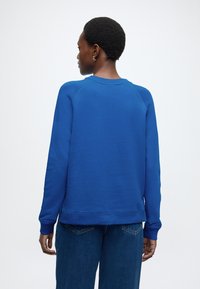 Kék pulóver hosszú ujjakkal, bordázott mandzsettákkal és kerek nyakkivágással. Vékony, sima anyagból készült, lazább szabással, sötét farmernadrággal párosítva.