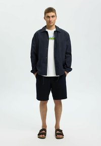 Niebieska lekka kurtka, biała koszulka z grafiką, ciemne szorty i brązowe sandały. Posiada zapięcie na guziki, szorty ze sznurkiem oraz teksturowany materiał.