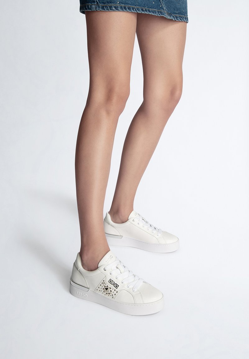 Sneaker bianchi in pelle con pannello laterale testurizzato e borchie, lacci piatti e suola spessa, indossati da un modello.
