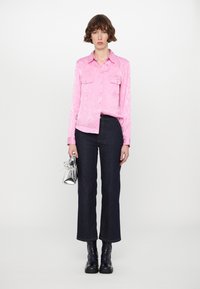 Pink, textured long-sleeve shirt with a collar and two chest pockets, paired with dark blue jeans and black ankle boots, holding a silver bag.