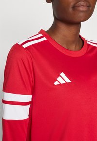 Maglietta sportiva rossa con colletto girocollo, maniche corte e strisce bianche. Presenta un logo Adidas bianco sul davanti. Tessuto liscio.