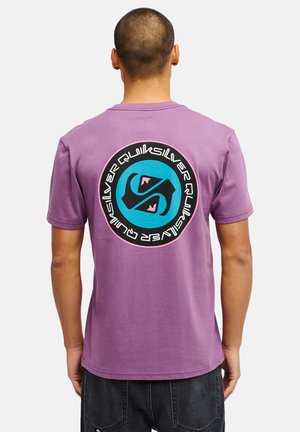 Homme aux cheveux courts portant un t-shirt violet avec un grand logo rond Quiksilver noir, bleu et rose dans le dos et un pantalon en jean noir.