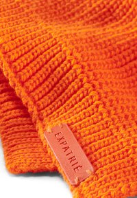 Lys orange strikket beanie med ribbet tekstur. Udstyret med et rektangulært mærke præget med "EXPATRIÉ" i en kontrasterende orange nuance.