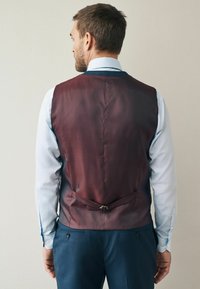 Mann mit blauer Hemd, marineblauen Hosen und burgunderrotem Westen, der von einer schlichten hellen Wand weg schaut.