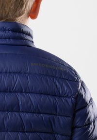 Giacca imbottita blu navy con motivo trapuntato orizzontale, dotata di colletto alto e dettagli con la scritta "SWEDEMOUNT" sul retro.