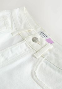 Shorts en denim blanc avec fermeture à bouton métallique, coutures visibles, passants pour ceinture et une étiquette de vêtement partiellement visible portant l'inscription « Hourglass Festival Short ».