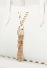 Borsa bianca con superficie liscia, caratterizzata da un accento a forma di V in oro e una frangia a catena attaccata a una chiusura in oro con incisione "V".
