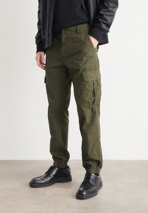 Pantaloni cargo verde oliva con polsini a gomito elasticizzati, tasche laterali e una vestibilità morbida, abbinati a scarpe nere stringate.