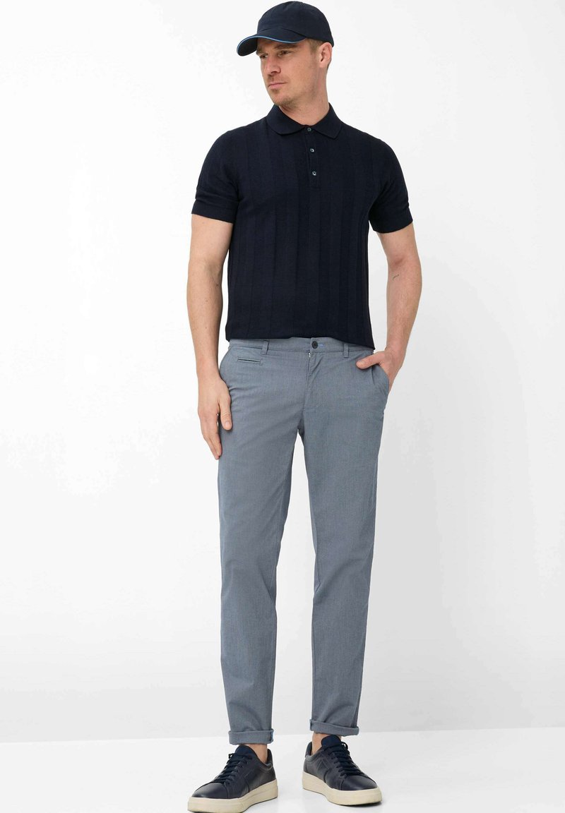 Man met een marineblauwe pet, zwart poloshirt met korte mouwen, lichtgrijze opgerolde broek en zwarte sneakers met witte zolen, staand met handen in de zakken.