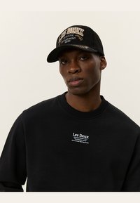Sudadera negra con cuello redondo y logo blanco "Les Deux". Gorra de pana negra con texto bordado y visera curvada.