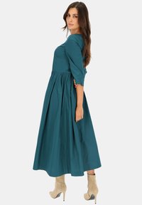 Turkusowa sukienka do kolan z rękawami o długości trzech czwartych, zebranym w pasie i zwiewnym dołem. Ma okrągły dekolt i gładką teksturę materiału.