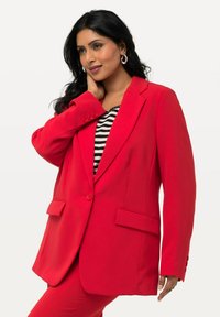 Blazer rosso con vestibilità sartoriale, chiusura a un bottone, due tasche frontali e rever a punte, abbinato a una maglietta a righe sotto.