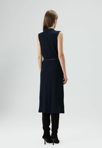 Marineblauwe mouwloze midi-jurk met een getailleerde pasvorm, voorzien van een ronde halslijn en een gladde textuur. Gecombineerd met kniehoge donkere laarzen.