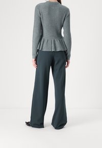 Femme portant un pull à basque côtelé gris et un pantalon large foncé, debout de dos sur un fond blanc uni.