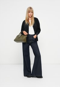 Czarna zamszowa kurtka zapinana na guziki, biała bluzka, szerokie ciemne dżinsy, trzymająca zieloną torebkę, na jednolitym białym tle.