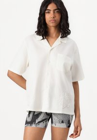 Camicia bianca a maniche corte con colletto, bottoni frontali e una tasca sul petto; presenta dettagli floreali ricamati vicino all'orlo.
