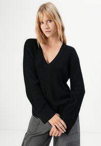 Woman with blonde hair wearing a black V-neck knit sweater and grey cargo pants, standing with hands clasped against white background.