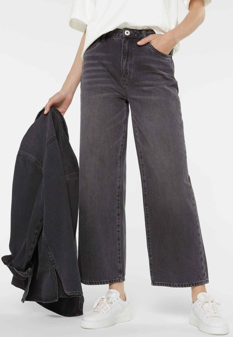 Wide-leg, dark grey jeans made of denim with a high waist, front pockets, and a button closure; paired with a matching jacket and white sneakers.