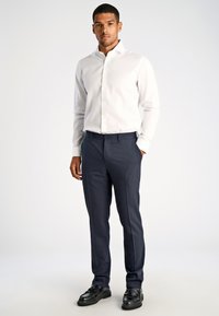 Camicia bianca con bottoni e maniche lunghe, abbinata a pantaloni sartoriali blu navy. Scarpe nere completano il look. Design semplice e pulito.