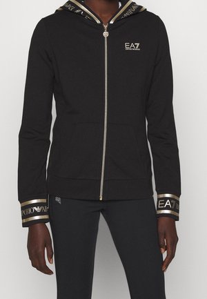 Personne portant un sweat à capuche noir zippé avec des poignets et une capuche bordés d'or, avec le logo EA7 Emporio Armani sur la poitrine et les manches.