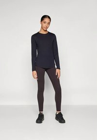 Vrouw die tegen een effen muur staat, gekleed in een zwart langemouwen shirt, donkere leggings en zwarte sneakers, met een iets voorwaartse blik en een ontspannen houding.