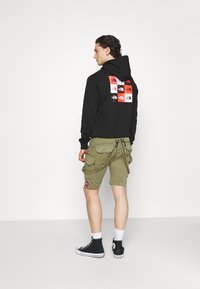 Hanorac negru cu imprimeu logo multicolor pe spate, asortat cu pantaloni scurți cargo verde măsliniu și adidași negri cu glezna înaltă.