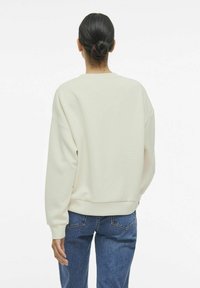Sudadera color crema con corte relajado, puños y dobladillo acanalados. Combinada con jeans de mezclilla azul. Textura suave, diseño minimalista.
